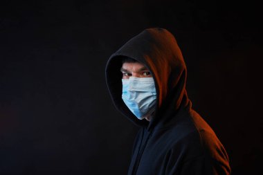 Siyah kapüşonlu, siyah kapüşonlu, sağlık maskeli, siyah arka planda renkli ışıkları olan genç moda adamı portresi.