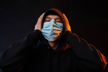 Siyah kapüşonlu, siyah kapüşonlu, sağlık maskeli, siyah arka planda renkli ışıkları olan genç moda adamı portresi.
