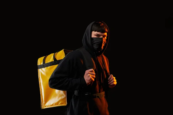 young delivery man in white clothes in rubber mask with a yellow backpack on a black background with orange light