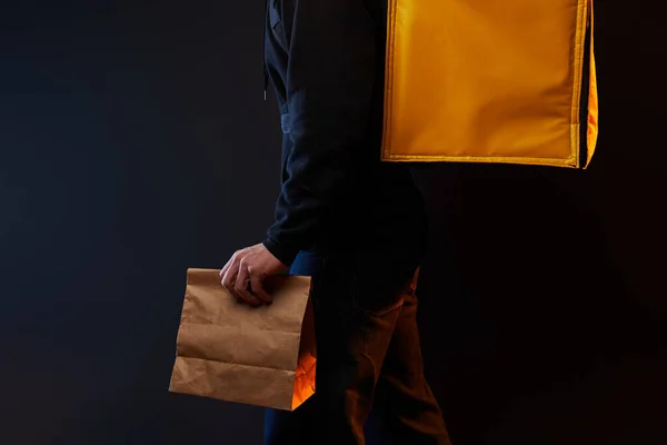 young delivery man in white clothes in rubber mask with a yellow backpack and on hand paper bag on a black background with orange light