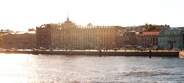 Panorama of the morning sun illuminates the city street and houses standing on the embankment of the river, passers-by go to work and walk, view from the opposite shore. — Stock Photo, Image