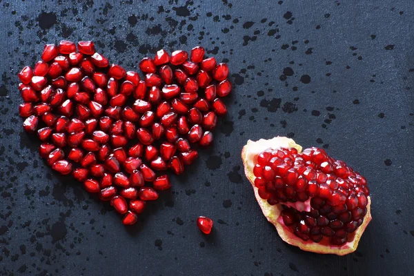 Pomegranate seeds are stacked in the form of a heart on a black background, sprinkled with water, next to a quarter of a pomegranate. — Stock Photo, Image