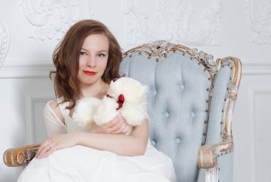 Beyaz elbiseli esmer kız derin bir koltukta oturur ve bir oyuncak ayı tutar