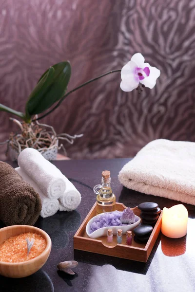 Conjunto para procedimentos de spa na mesa, toalhas terry, sais de banho, pedras de massagem e bio-óleo. Orquídea florida no fundo, atmosfera quente . — Fotografia de Stock