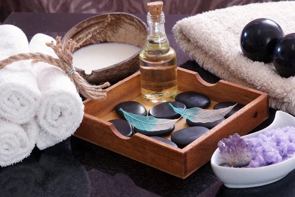 Weiße Handtücher, gestrickt neben dem Set für die Massage aus bianischen Steinen, Aromaöl und violettem Salz, in einer Holzkiste ein Satz heißer Steine zur Entspannung und für eine Wellness-Massage. — Stockfoto