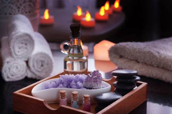 Conjunto de procedimentos de spa - pedras de massagem, óleo, sal do mar de ametista violeta e natural encontra-se em uma caixa. Toalhas úmidas encontram-se em uma mesa de granito preto natural. Velas elétricas queimam no fundo . — Fotografia de Stock