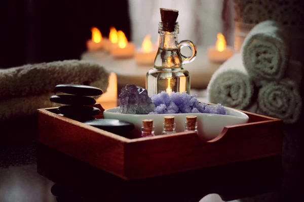 Conjunto para procedimentos de spa de pedras para massagem, óleo, sal marinho de violeta encontra-se em uma caixa de madeira. Toalhas úmidas encontram-se em uma mesa de granito preto natural.Velas elétricas queimam . — Fotografia de Stock