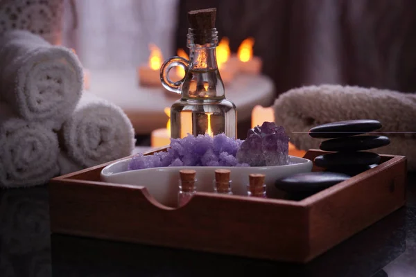 Conjunto para procedimentos de spa de pedras para massagem, óleo, sal marinho de violeta encontra-se em uma caixa de madeira. Toalhas úmidas estão em uma mesa de granito preto natural.Velas elétricas queimam no fundo . — Fotografia de Stock