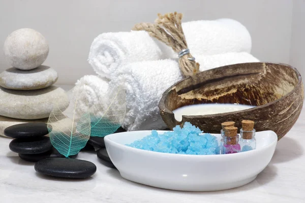 Un conjunto de tratamientos de spa con leche de coco, piedras calientes y sal de baño azul se encuentra en una encimera de mármol blanco . — Foto de Stock