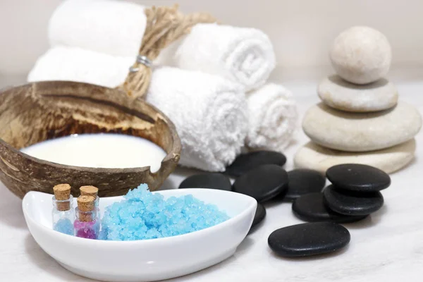 Un conjunto de tratamientos de spa con leche de coco, piedras calientes y sal de baño azul se encuentra en una mesa de mármol blanco . — Foto de Stock