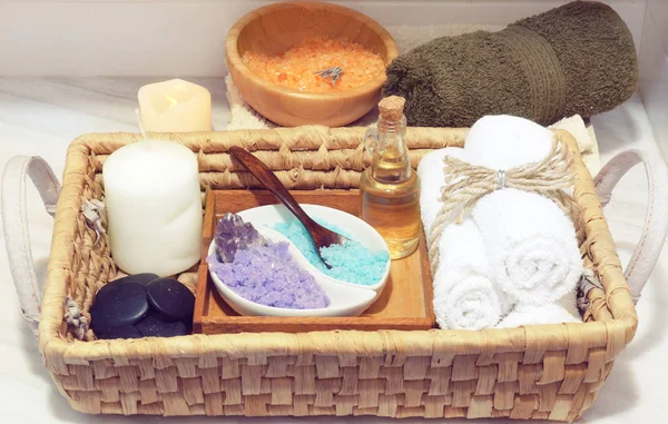 Korg med en uppsättning för spa-behandlingar, flerfärgad salt, aromatisk olja, stenar, ljus och mjuka handdukar, bredvid ett vitt bord träskål med havssalt — Stockfoto
