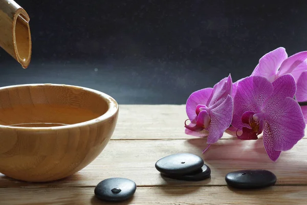 Kom, giet die transparant water uit de bamboe stam, orchideebloemen en stenen voor een hete massage op een houten tafel — Stockfoto