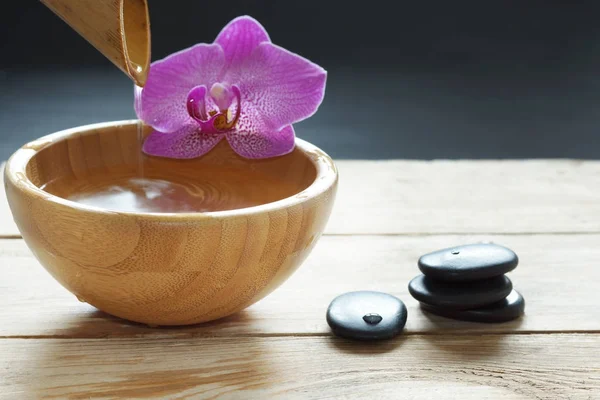 Kom, giet die transparant water uit bamboe, orchideebloemen en stenen voor een hete massage op een houten tafel — Stockfoto