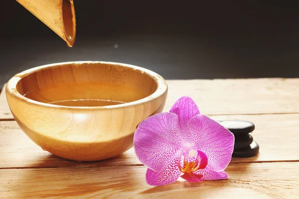 Kom, die giet water uit de bamboe stam, orchideebloemen en stenen voor een hete massage op een houten tafel — Stockfoto