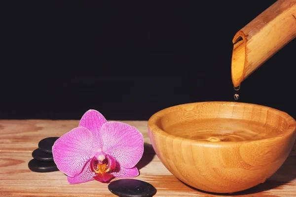 Water drupt van de bamboe in een kom op een houten tafel, naast de spa behandeling stenen en de orchideebloem, kopie ruimte voor uw tekst — Stockfoto