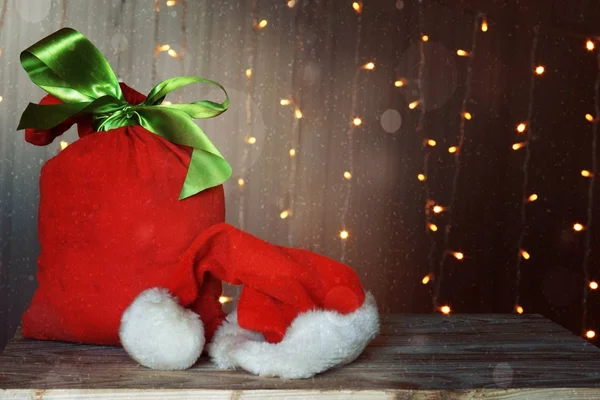 Рождественская открытка с красной сумкой, полной подарков и шляпой Санта Клауса. Светящиеся гирлянды на заднем плане . — стоковое фото