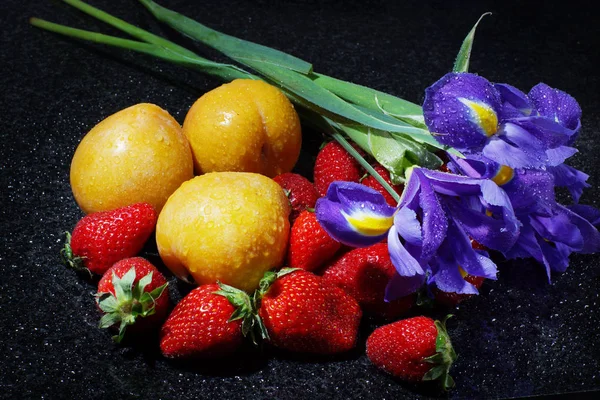 Natureza morta em uma mesa escura com íris, muitos morangos e ameixas amarelas com gotas de água — Fotografia de Stock