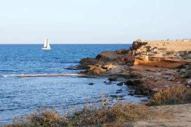 Bir uçurum kıyı taşla çerçevede bir denizde bir beyaz yelkenli yelken.