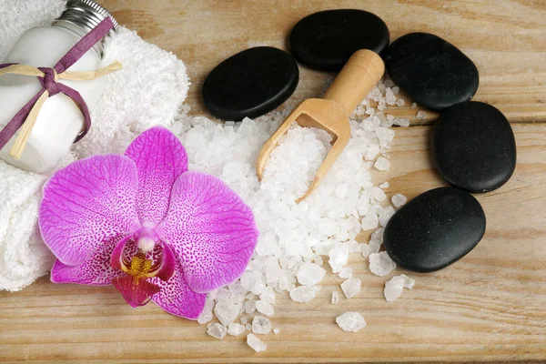 Conjunto para tratamentos de spa com loções para pele, flores de orquídeas, sal de banho e pedras pretas para uma massagem quente em um fundo de madeira — Fotografia de Stock