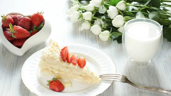 Un pedazo de pastel Napoleón, un tazón de fresas, un ramo de flores y un vaso de leche en una mesa por la mañana temprano — Foto de Stock