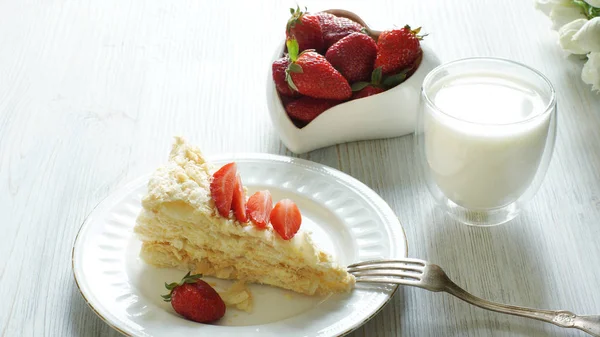 Un pedazo de pastel de Napoleón y un tazón en forma de corazón con una fresa roja, un vaso al lado de la leche, un bodegón ligero — Foto de Stock