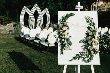 White board with lettering 'Welcome' and green leaves with white roses stands on the easel clipart