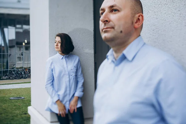 Man and woman in blue shirts and jeans stand apart before a grey building