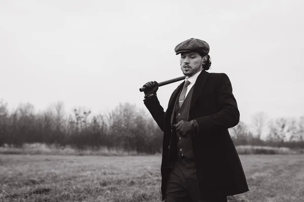 Fashionable photo of young attractive man wearing in stylish vintage suit. Man with tweed hat looking down and holding baton on back. Concept of fashion and vintage.