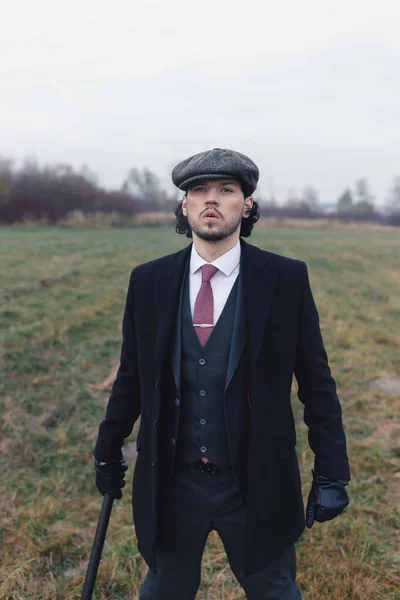 Portrait of attractive vintage man wearing in tweed blinder, black pants and coat, leather gloves and red tie. Male with curly dark hair looking at sky and holding black baton. Concept of fashion and 20`s.