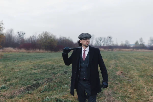 Attractive vintage man with curly dark hair, wearing in black coat, pants and red tie, holding baton on his shoulder and looking away. Fog, field and trees on background. Concept of fashion and vintage.