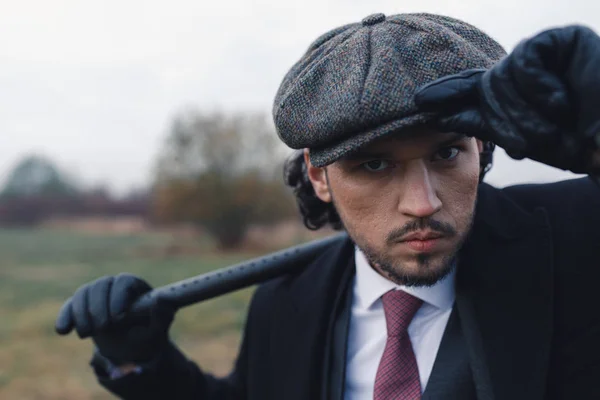 Caucasian handsome man in 20`s style correcting his tweed blinder, wearing in black leather gloves, red tie and black jacket. Concept of fashion and vintage.