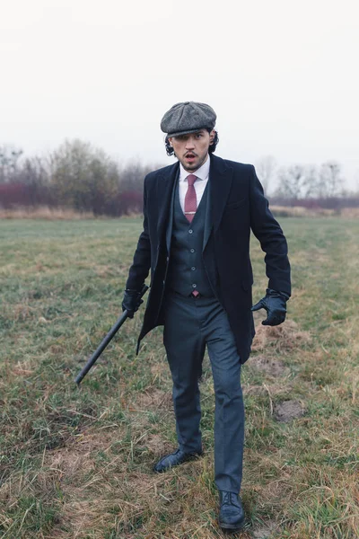 一个严肃的男人 身穿黑色衣服 头戴粗花呢帽 站在田野中间 看着相机 拿着警棍 时尚和复古的概念 免版税图库图片