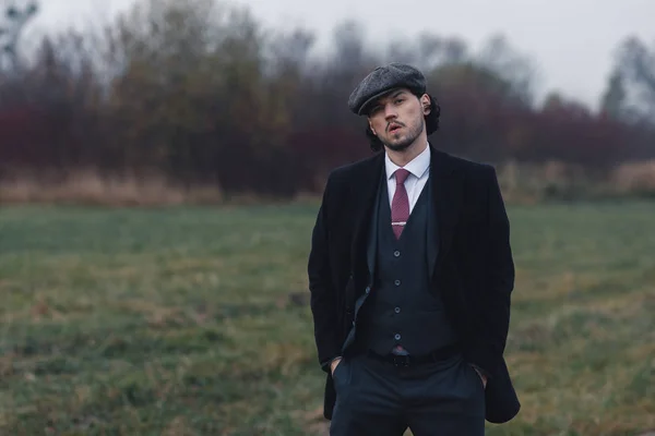 Adorable husband in stylish classical suit standing in autumt foggy forest. Man wearing in black clothes and looking at camera. Concept of fashion and classic.