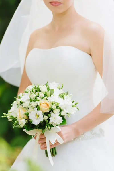 Bride in a white dress holds a beautiful bouquet of white flowers and greenery, decorated with silk ribbon — Stock Photo, Image