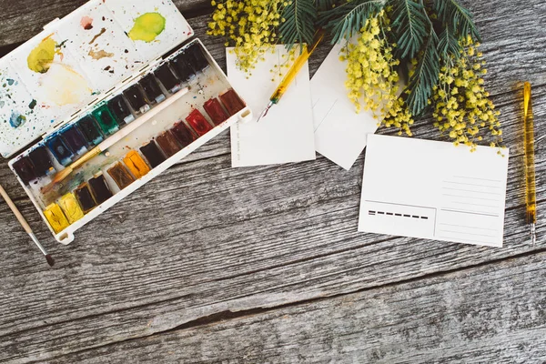 Espacio de trabajo. Marco floral con mimosas, acuarelas, pincel y postal vintage sobre fondo de madera . — Foto de Stock