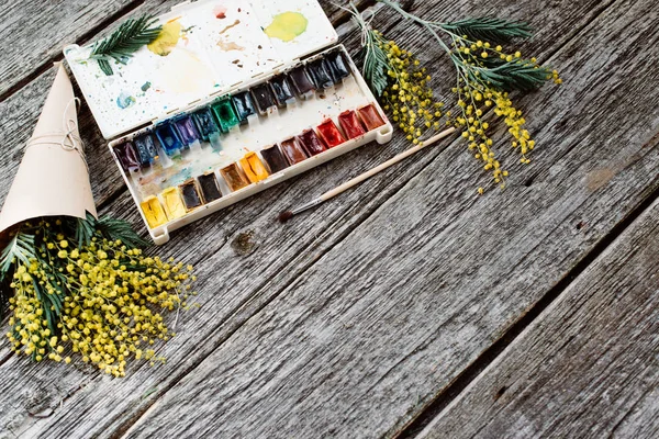 Espacio de trabajo. Marco floral con mimosas, acuarelas, pincel sobre fondo de madera . — Foto de Stock