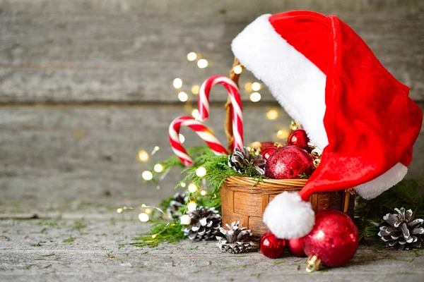 Holiday lesklé složení vánoční koš s míčky, hračky, sladkosti a Borové šišky, santa claus klobouk na vintage dřevěné pozadí — Stock fotografie