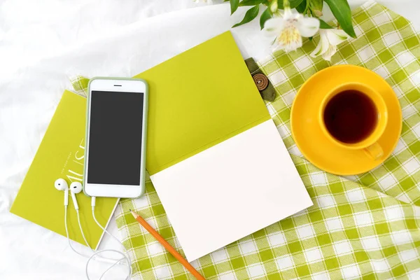 Telefon leżał płasko, żółty filiżankę herbaty i kwiaty na biały koc z zielone serwetki — Zdjęcie stockowe