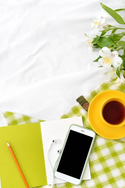 Teléfono plano, taza amarilla de té y flores en manta blanca con servilleta verde —  Fotos de Stock