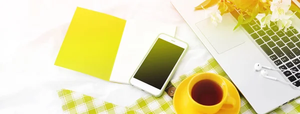 Banner largo Teléfono plano, taza amarilla de té, computadora portátil y flores en manta blanca con servilleta verde — Foto de Stock