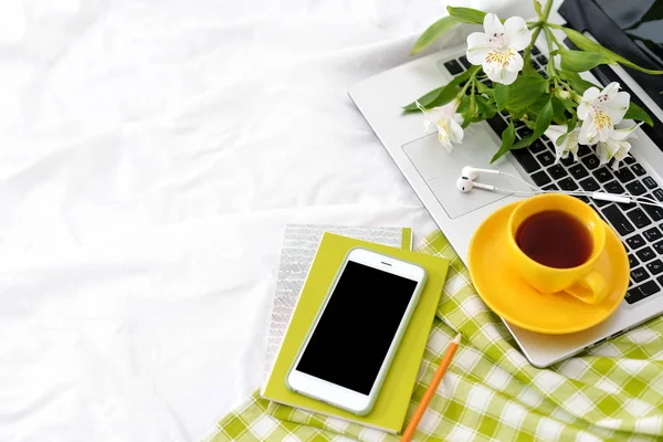 Плоский планшет, телефон, жовта чашка чаю, ноутбук і квіти на білій ковдрі з зеленою серветкою — стокове фото