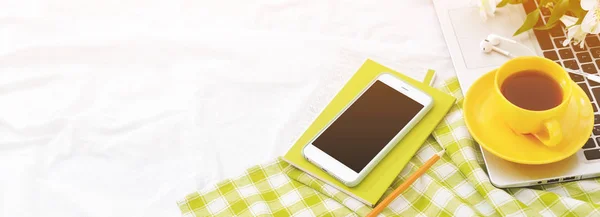 Long banner Flat lay phone, yellow cup of tea, laptop and flowers on white blanket with green napkin