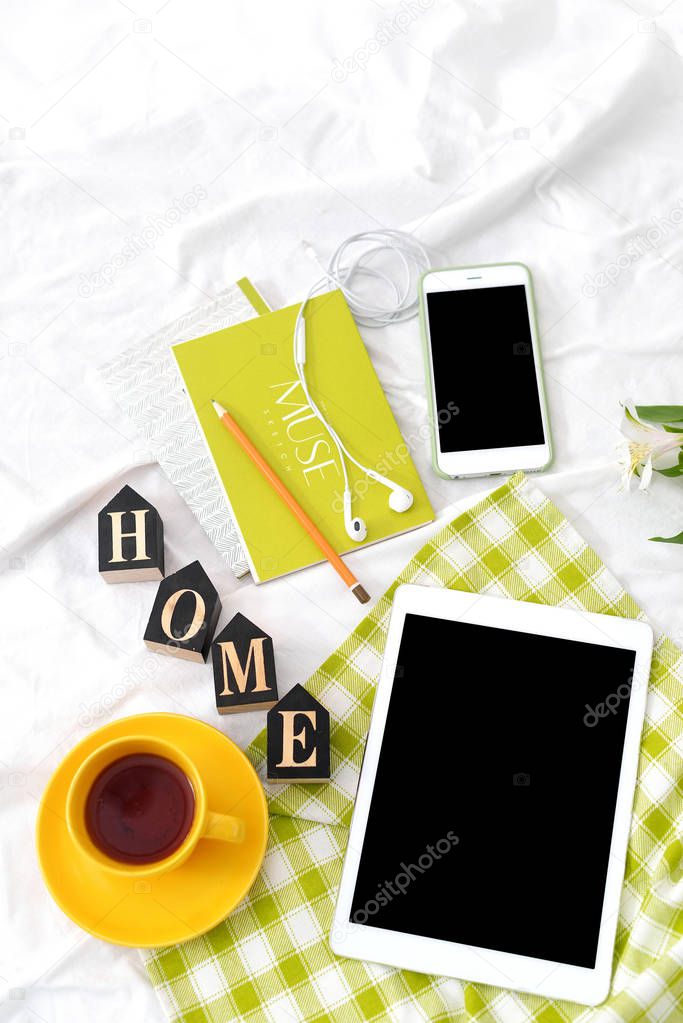 Flat lay tablet, phone, yellow cup of tea, laptop and flowers on white blanket with green napkin