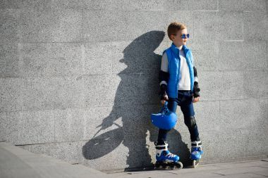 Satır içi tekerlekli paten ve açık skate Park tüm koruma ile gri duvarının yakınında duran genç çocuk