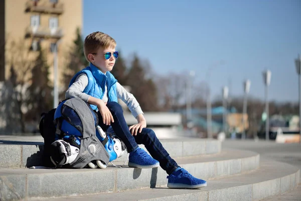 Giovane ragazzo seduto vicino al muro grigio con pattini a rotelle in linea e tutta la protezione allo skate park all'aperto — Foto Stock