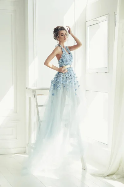 young beautiful lady in blue long romantic dress. fairytale morning light. trendy bridal hairstyle. white room.