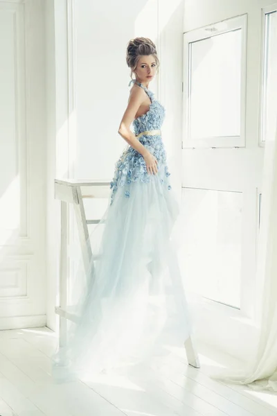 young beautiful lady in blue long romantic dress. fairytale morning light. trendy bridal hairstyle. white room.