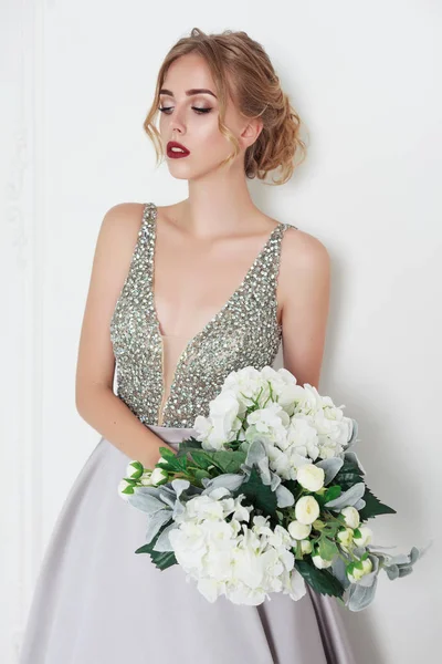 young beautiful blonde woman in silver dress. luxury bridal dress. lady with red lips and trendy hairstyle. lady on white background. bouquet with white flowers