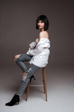 Kısa saçlı güzel bir esmer. Beyaz ceket ve kot pantolon giyen bir manken. Gri stüdyo arka planında poz veren günlük elbiseli bir bayan. Bar sandalyesinde manken. moda endüstrisi