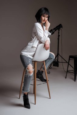 Kısa saçlı güzel bir esmer. Beyaz ceket ve kot pantolon giyen bir manken. Gri stüdyo arka planında poz veren günlük elbiseli bir bayan. Bar sandalyesinde manken. moda endüstrisi
